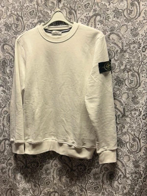 Vit sweatshirt från Stone Island - Säljer en stilren vit sweatshirt från Stone Island med klassisk rund hals och ribbade muddar. Tröjan har den ikoniska svarta logotypen med kompass på vänster ärm. Perfekt för dig som gillar streetwear och enkel design.
