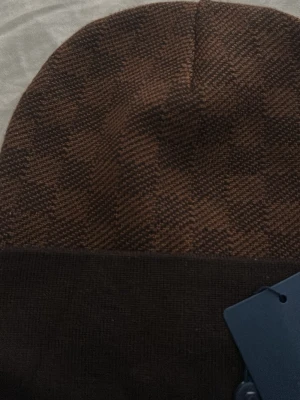 Brun rutig stickad mössa - Snygg brun stickad mössa med rutigt mönster och uppvikt kant. Perfekt accessoar för kyliga dagar och passar till de flesta outfits. Mjuk och skön att bära.