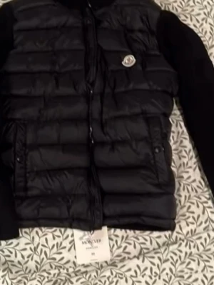 Svart Moncler cardigan  - Snygg svart dunjacka från Moncler med quiltad front och ärmar i mjukt material. Jackan har dragkedja framtill, två fickor med tryckknappar och Moncler-logga på bröstet. Perfekt för kalla vinterdagar och riktigt stilren look.