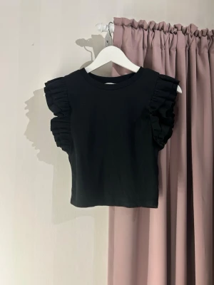 Svart croppad topp med volangärmar - Snygg svart croppad topp med volangdetaljer på ärmarna. Toppen har rund halsringning och är tillverkad i ett mjukt, ribbat material som ger en skön passform. Perfekt för dig som vill ha en trendig och stilren look.