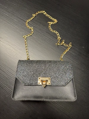Svart glittrig väska med kedja - Snygg svart axelväska med glittrigt lock och guldfärgad kedja. Väskan har en rektangulär form och ett dekorativt guldfärgat lås framtill. Materialet är en mix av glittrig textil och slät konstläder. Perfekt för att lyfta din outfit med lite extra bling.