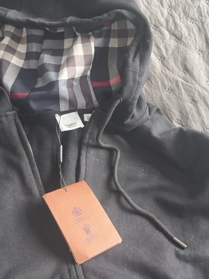 Svart Burberry hoodie med rutigt foder - Säljer en svart hoodie från Burberry med klassiskt rutigt foder i huvan. Hoodien har dragkedja framtill, snörning i huvan och diskret Burberry-logga på bröstet. Perfekt för dig som vill ha en stilren och lyxig hoodie med ikonisk design.