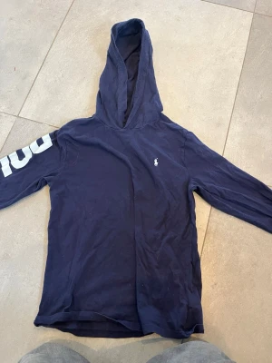 Mörkblå hoodie från Polo Ralph Lauren - Mörkblå hoodie från Polo Ralph Lauren med klassisk logga broderad. Hoodien har lång ärm och en rymlig huva. Tillverkad i mjuk bomull för en avslappnad och bekväm stil.