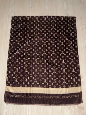 Brun Louis Vuitton sjal med monogram - Snygg brun sjal från Louis Vuitton med klassiskt monogrammönster i beige och bred kant med logotyp. Tillverkad i ett mjukt, glansigt material som känns lyxigt och har fransad kant. Perfekt accessoar för att lyfta din outfit.