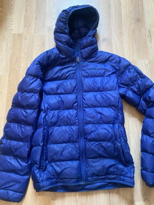 Mörkblå dunjacka från Peak Performance - Snygg blå dunjacka från Peak Performance med huva och dragkedja framtill. Jackan har två sidofickor med dragkedja och quiltad design. Perfekt för kalla vinterdagar och riktigt skön att bära. Klassisk logga broderad på bröstet.