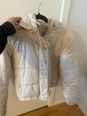 Beige pufferjacka med huva - Snygg beige pufferjacka med huva och dragkedja samt tryckknappar framtill. Jackan har quiltad design och två sidofickor. Perfekt för kalla dagar och ger en trendig look. Materialet är troligtvis polyester.