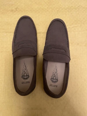 Bruna loafers från Burton - Snygga bruna loafers från Burton i mockaimitation. Klassisk penny loafer-design med rundad tå och diskret söm längs kanten. Insidan har tryckt logga och skon har en stilren, minimalistisk look som passar till många outfits. Färgen är grön/brun