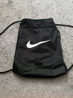 Svart gympåse från Nike - Snygg svart gympåse från Nike med klassisk vit swoosh-logga på framsidan. Väskan har dragskostängning, en dragkedjeficka framtill och är tillverkad i 100% polyester. Perfekt för träning eller skola.