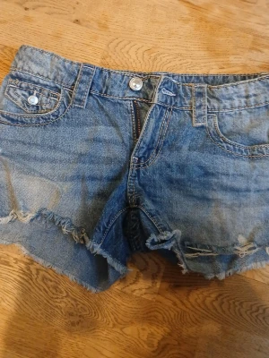Blå jeansshorts med fransar - Säljer ett par blå jeansshorts med råa fransade kanter och klassisk femficksdesign. Shortsen har knappgylf och bälteshällor, samt en ficka med lock och knapp bak. Perfekta för sommaren och ger en avslappnad look.