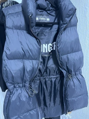 Svart dunväst från Stronger - Snygg svart dunväst från Stronger med hög krage och dragkedja framtill. Västen har justerbar midja med snörning och quiltad design som ger en puffig look. Perfekt för lager på lager och streetstyle.