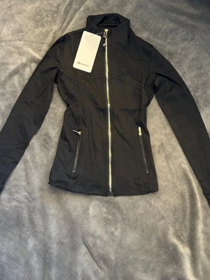 Svart träningsjacka från Lululemon - Slimmad svart träningsjacka från Lululemon med hög krage, hel dragkedja framtill och två dragkedjeförsedda fickor. Tillverkad i stretchigt funktionsmaterial som andas. Snygga sömdetaljer och tumhål vid ärmsluten för extra komfort.