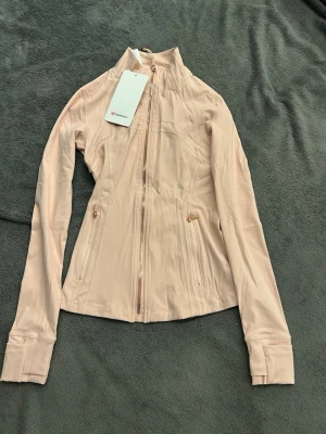 Ljusrosa träningsjacka Lululemon - Snygg ljusrosa träningsjacka från Lululemon med figurnära passform, hög krage och hel dragkedja framtill. Jackan har två fickor med dragkedja och långa ärmar med tumhål. Tillverkad i ett stretchigt funktionsmaterial som andas.