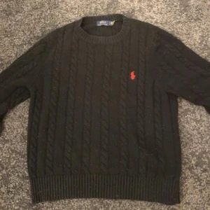 Svart kabelstickad tröja Polo Ralph Lauren - Kabelstickad svart tröja från Polo Ralph Lauren med rund halsringning och röd broderad logga på bröstet. Klassisk modell med ribbade muddar vid ärmslut och nederkant. Perfekt för dig som gillar stilrena och tidlösa plagg.