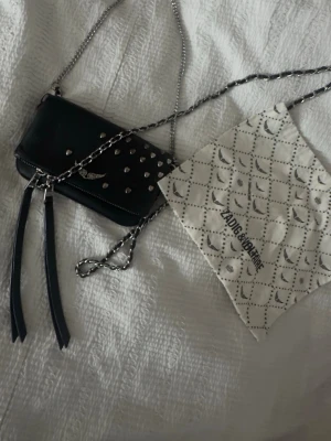 Svart axelväska Zadig & Voltaire - Säljer en svart axelväska från Zadig & Voltaire i läder med silverfärgade nitar och kedja. Väskan har dekorativa tofsar, ett emblem med vingar och magnetlås. Kommer med original dustbag. Perfekt för dig som gillar edgy detaljer och stilren design.
