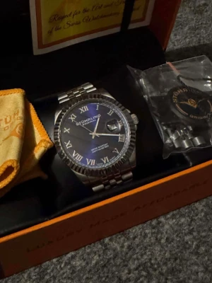 Stührling klocka - Snygg klocka från Stührling med blå urtavla, romerska siffror och datumvisning. Armbandet är i silverfärgad metall och har klassisk länkdesign. Kommer i originalask med extra länkar och putsduk.