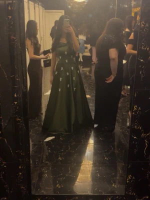 Grön axelbandslös långklänning - Elegant grön axelbandslös långklänning med utsvängd kjol och dekorativa blommor framtill. Klänningen har en vacker offshoulder-design och passar perfekt till speciella tillfällen. Materialet har en lätt glansig finish.