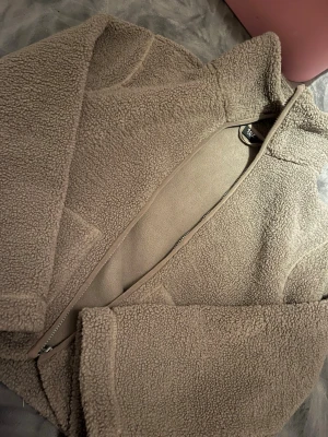 Beige teddyjacka från lager 157 - Mysig beige teddyjacka från Lager 157 med dragkedja framtill och två stora fickor. Jackan har en hög krage och är gjord i fluffigt teddy-material som håller dig varm. Perfekt för kyliga dagar och ger en trendig look.