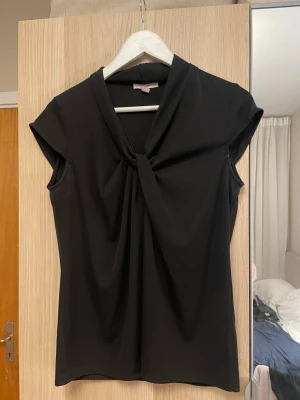 Svart omlottopp från R.O.B. - Stilren svart omlottopp från R.O.B. med korta ärmar och snygg twistad detalj vid halsringningen. Toppen har en mjuk och följsam passform som ger en elegant look.