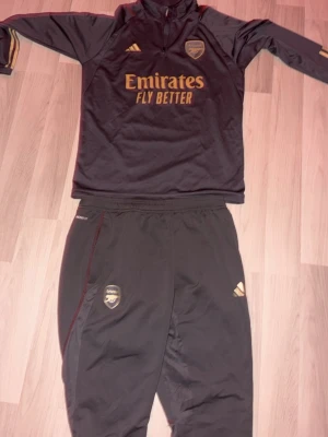 Arsenal träningsset svart/guld Adidas - Snyggt svart träningsset från Adidas och Arsenal med guldiga detaljer och loggor. Tröjan har halv dragkedja och byxorna har resår i midjan. Både tröja och byxor har Arsenal-emblem och Adidas-logga. Perfekt för fotbollsträning eller chill. Skick 10/10 inte alls använd.