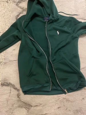 Mörkgrön hoodie från Polo Ralph Lauren - Snygg mörkgrön hoodie från Polo Ralph Lauren med dragkedja, huva och fickor framtill. Slim fit-modell i stretchmesh-material som ger skön passform. Klassisk vit logga broderad på bröstet. Perfekt för en avslappnad och stilren look.