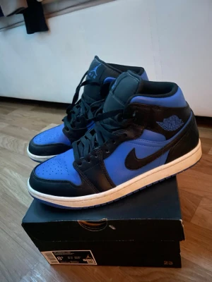 Air Jordan 1 Mid Svart/Blå - Säljer ett par Air Jordan 1 Mid sneakers i svart och blått med vita sulor. Klassisk siluett med svart Swoosh och blåa detaljer på tå, häl och logga vid ankeln. Tillverkade i läder och textil, perfekta för dig som gillar streetwear och sneakers med retrovibe.