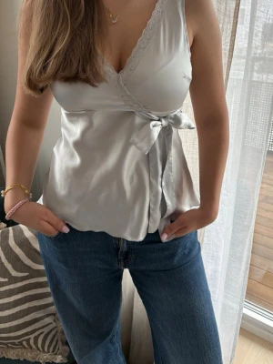 Silvrig omlottopp med spetsdetaljer - Elegant topp från Ellos i silver/ljusblå satin🌟  Skickas snabbt!📦  Modellen har omlottdesign med V-ringning, vacker spetskant samt knytband i sidan som ger en feminin passform. Lätt utsvängd nedtill.  Storlek: 38 Skick: Använd, i fint skick.