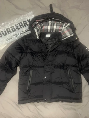 Svart pufferjacka från Burberry - Snygg svart pufferjacka från Burberry med klassiskt rutigt foder i huvan. Jackan har dragkedja och tryckknappar framtill, två snedställda fickor med dragkedja och logga på ärmen. Perfekt för kalla vinterdagar och riktigt trendig look. Kan ta av ärmarna för att få en väst, utmärkt för olika årstider.