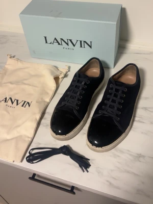 Svarta sneakers från Lanvin - Snygga svarta sneakers från Lanvin med ovandel i mocka och lackad tå. Skorna har vita sulor och klassisk snörning. Skicket är 6/10. Perfekta för dig som gillar stilrena och exklusiva detaljer. Allt og medföljer.