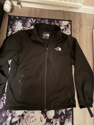 Svart vindjacka från The North Face - Svart vindjacka från The North Face med dragkedja framtill, två sidofickor med dragkedja och en bröstficka. Jackan har hög krage, justerbara muddar och är tillverkad i ett lätt och slitstarkt material. Perfekt för blåsiga dagar.