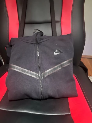 Svart Nike zip hoodie med huva - Säljer en svart zip hoodie från Nike med huva och dragkedja framtill. Hoodien har diskreta svarta detaljer i form av två diagonala linjer över bröstet och en liten Nike-logga på vänster sida. Perfekt för en sportig och avslappnad stil.