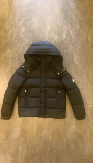 Svart Moncler vezere dunjacka med huva - Snygg svart dunjacka från Moncler med avtagbar huva, dragkedja och knappar framtill. Jackan har klassisk puffer-design, två fickor med dragkedja och logotyp på ärmen. Perfekt för kalla dagar och riktigt stilren look. Pris kan diskuteras 