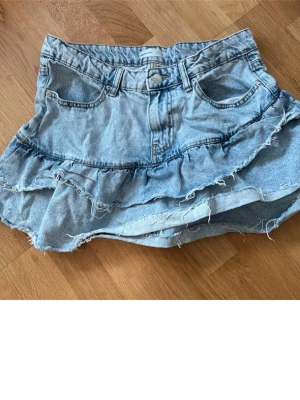 Blå jeanskjol med volang - Snygg kort jeanskjol i ljusblå denim med volangdetaljer och råa, slitna kanter. Kjolen har klassiska fickor fram och bak samt en avslappnad, trendig look. Perfekt för dig som gillar en cool och avslappnad stil.