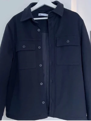 Overshirt från Zara - Stilren svart overshirt från Zara med klassisk krage, knäppning framtill och två bröstfickor med lock och knapp. Overshirten har en lätt vadderad insida som ger extra komfort och värme. Perfekt att bära som lager över en hoodie eller t-shirt.