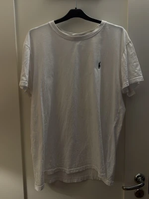 Vit t-shirt från Ralph Lauren - Klassisk vit t-shirt från Ralph Lauren med liten broderad logga på bröstet. T-shirten har rund halsringning och korta ärmar. Tillverkad i mjuk bomull för en skön och avslappnad stil. Perfekt basplagg till garderoben.