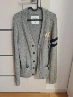 Grå kofta från Abercrombie & Fitch - Mysig grå kofta från Abercrombie & Fitch med knappar framtill, två fickor och ribbade muddar. Broderad AF-logga på bröstet och två mörkblå ränder på ena ärmen ger en sportig touch. Perfekt för lager på lager och chill stil.