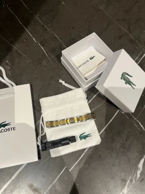Lacoste guldfärgad armband - Snygg armband från Lacoste med guldfärgad och silvrig metall-länk. Kommer i originalförpackning med påse och verktyg för att justera länken. Perfekt accessoar för dig som vill ha en stilren och modern look.