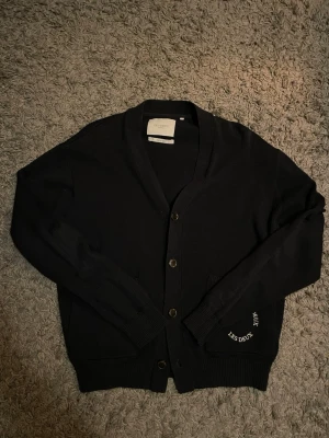 Mörkblå cardigan från Les Deux - Stilren mörkblå cardigan från Les Deux med knäppning framtill och diskret broderad logga vid nederkanten. Tröjan har v-ringad hals och ribbade muddar. Perfekt för lager på lager och enkel att matcha med olika outfits.