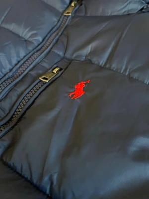 Mörkblå pufferjacka från Polo Ralph Lauren - Snygg mörkblå pufferjacka från Polo Ralph Lauren med röd broderad logga på bröstet. Jackan har huva, två dragkedjeförsedda fickor och quiltad design. Perfekt för kalla dagar och riktigt skön att bära. Materialet är glansigt och ger en modern look.