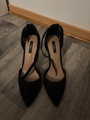 Svarta pumps med remmar från ONLY - Snygga svarta pumps från ONLY med spetsig tå och smala remmar över foten. Skorna har hög klack och justerbart spänne vid ankeln. Materialet är mocka. Använda en gång!