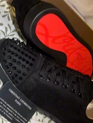 Svarta spikes sneakers Louboutin - Säljer ett par svarta sneakers från Christian Louboutin med mattsvarta nitar på tån och den ikoniska röda sulan. Skorna har svarta skosnören och är tillverkade i mocka och läder. Perfekta för dig som vill sticka ut med en edgy och lyxig stil.