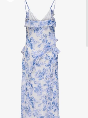Blommig långklänning med volanger -  Långklänning med blått blommönster på vit botten. Klänningen har tunna axelband, v-ringning och volangdetaljer framtill och längs sidorna. Perfekt för sommarens alla tillfällen och ger en fräsch och feminin look.