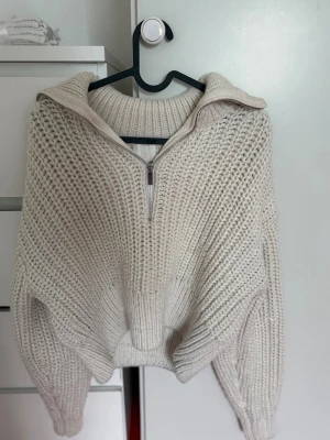 Beige stickad tröja med halv dragkedja - Säljer en beige stickad tröja med grov struktur och halv dragkedja framtill. Tröjan har hög krage, långa ärmar och ribbade muddar. Perfekt för lager på lager och en avslappnad stil.