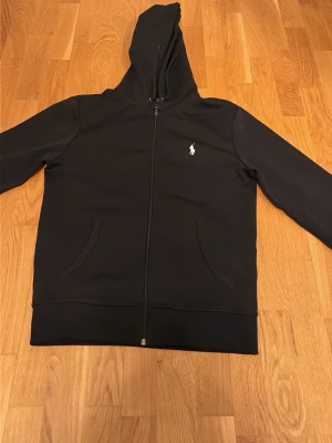 Svart hoodie från Polo Ralph Lauren - Svart hoodie med dragkedja från Polo Ralph Lauren. Klassisk design med vit broderad logga på bröstet, två fickor framtill och huva. Perfekt för en avslappnad och stilren look. Materialet känns mjukt och bekvämt mot huden. Den har ett litet jack i tyget där back, annars har den inga defekter 