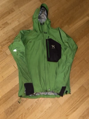 Grön Haglöfs vindjacka Gore-Tex - Snygg grön vindjacka från Haglöfs med svart bröstficka och huva. Tillverkad i Gore-Tex Active Shell som gör den både vattentät och andas. Jackan har dragkedja framtill, justerbar nederkant och muddar vid ärmsluten. Perfekt för uteliv och äventyr. finns några fläckar och pris är inte hugget i sten, skriv om ni vill ha fler bilder eller är intresserade