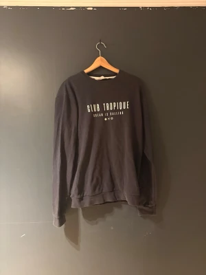 Svart sweatshirt Club Tropique - Cool svart sweatshirt från Club Tropique med vit text på bröstet. Tröjan har rund halsringning, långa ärmar och ribbade muddar vid ärmslut och nederkant. Perfekt för en avslappnad och trendig look.
