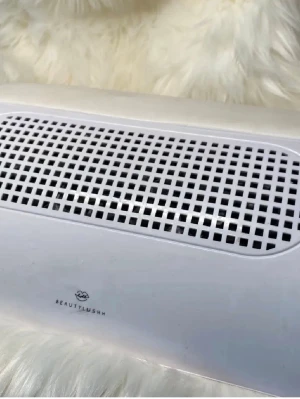 Nageldammsugare från Beautylushh - Nageldammsugare i vit plast med mjuk, vadderad handledskudde. Perfekt för att suga upp damm vid nagelfilning och ger en ren arbetsyta. Modern design med rutmönstrat galler och stilren finish. Passar både hemma och i salong.