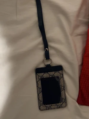 Gucci korthållare med halsband - Snygg korthållare från Gucci med halsband i svart läder och beige canvas med GG-monogram. Perfekt för att bära ID eller kort runt halsen. Klassisk design med transparent fönster på framsidan och detaljer i silverfärgad metall.