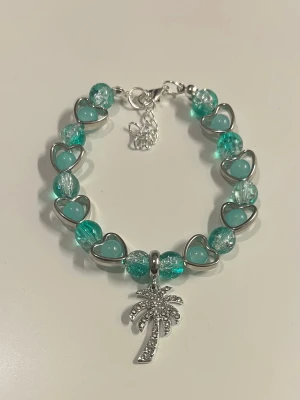 Armband med en palm berlock - Snyggt armband med turkosa och genomskinliga pärlor samt hjärtformade detaljer i silverfärg. Armbandet har en berlock i form av en glittrig palm. 