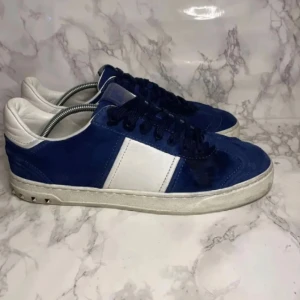 Valentino flycrew - Snygga sneakers från Valentino i blå mocka och vitt läder. Klassisk låg modell med vita detaljer och svarta skosnören. Yttersulan är vit med unika mönster under och små nitar på hälen. Perfekta för dig som vill ha en stilren men ändå cool look.