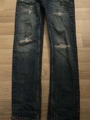 Blå straight fit jeans från Replay - Säljer ett par blå straight fit jeans från Replay med slitna detaljer och snygg tvätt. Jeansen har klassisk femficksmodell och raka ben. Perfekta för en avslappnad och trendig look.